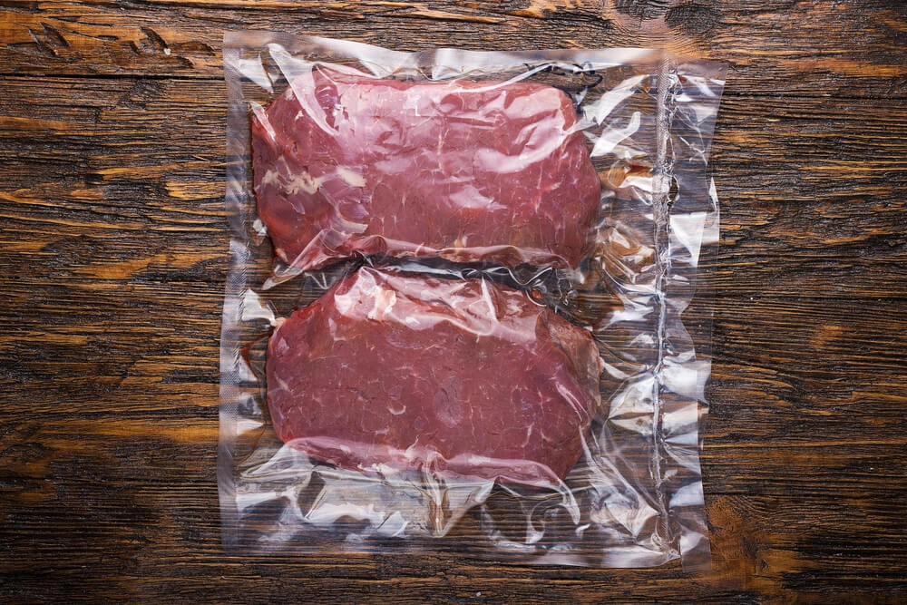 Auf dem Bild sieht man zwei Stücke Fleisch in einem glatten Vakuumbeutel eingeschweißt auf einer Holzplatte.